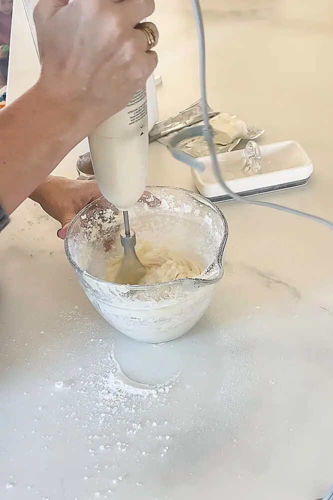 Hands holding an electric mixer while preparing cream cheese frosting in a glass measuring cup.