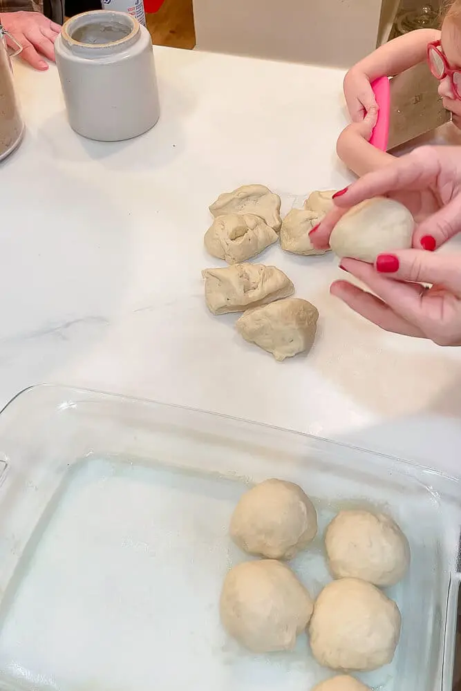Hands shaping small dough balls and placing them into a greased glass dish, getting them ready to bake into dinner rolls.