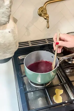 A person stirring raspberry gelatin in a saucepan on the stove, preparing the topping for the raspberry pretzel pie.