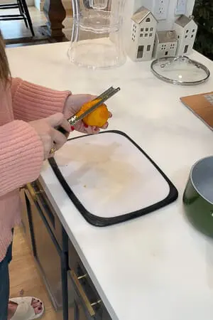 A person in a pink sweater zesting an orange over a small cutting board, preparing ingredients for cranberry sauce.
