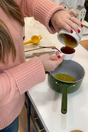A person pouring honey from a small cup into a green saucepan, with various kitchen tools and ingredients nearby.