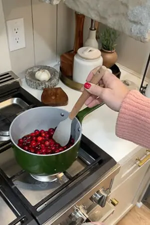 A person stirring fresh cranberries in a green saucepan on the stove, as the ingredients for cranberry sauce cook together.