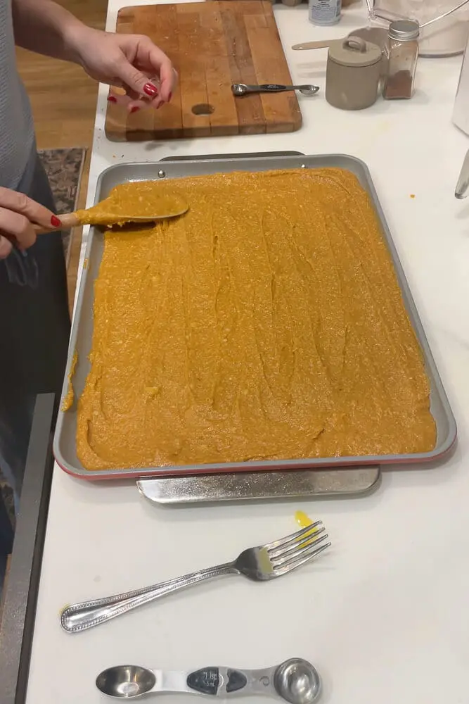 A person spreading pumpkin cake batter evenly onto a baking tray with a wooden spoon.