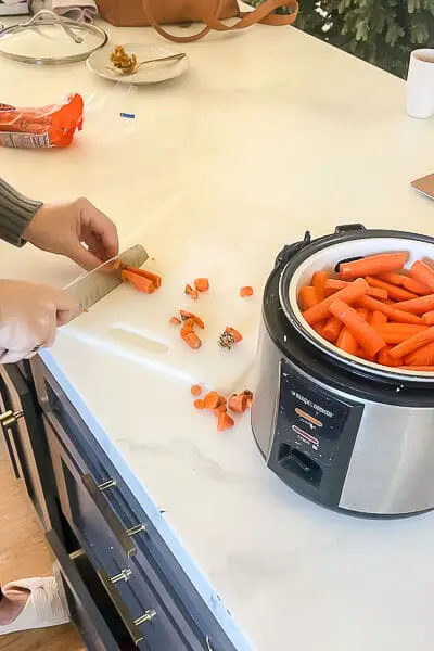A person chopping fresh carrots and adding them into a slow cooker for steaming.