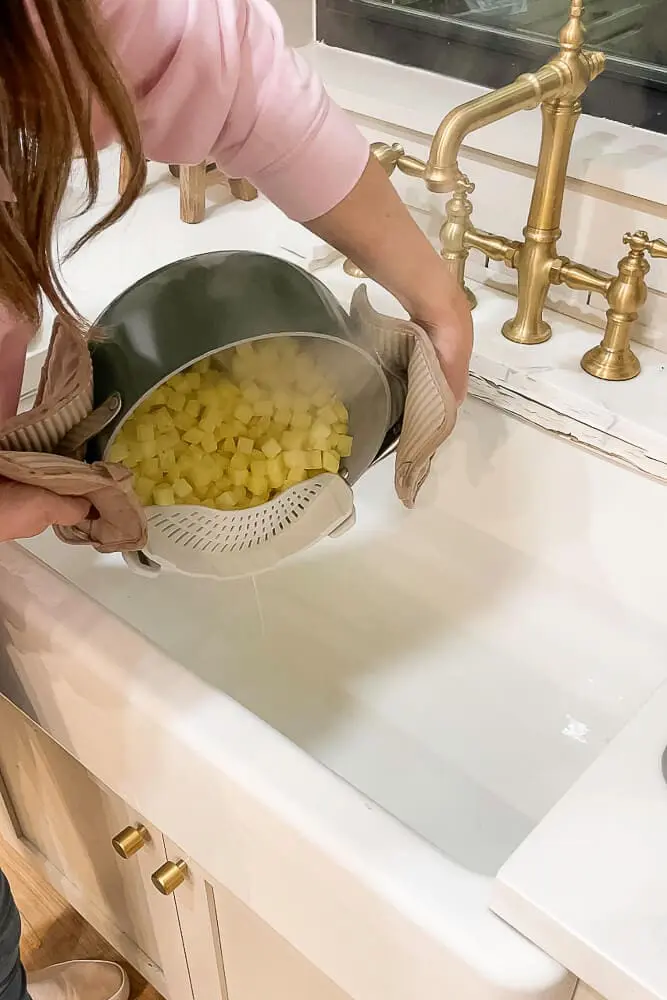 A person draining boiled diced potatoes into a colander over a sink, preparing for garlic mashed potatoes