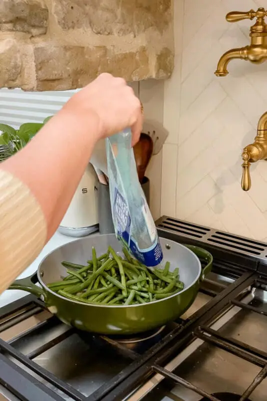 A hand pouring a bag of fresh green beans into a green pan on the stove.