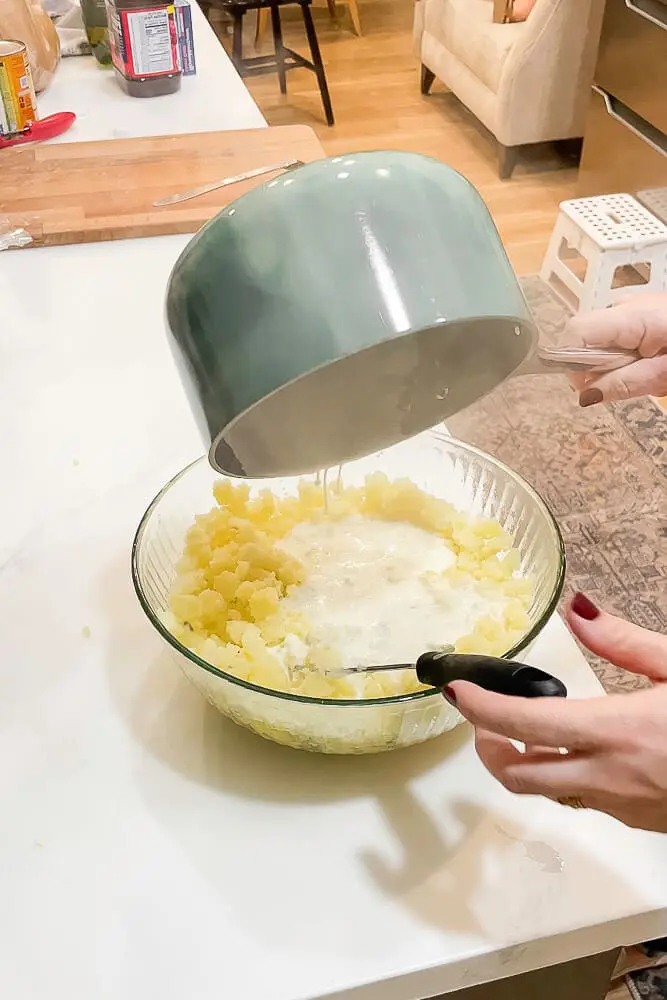 Warm cream being poured over cooked potatoes in a glass mixing bowl, ready to be mashed into garlic mashed potatoes.