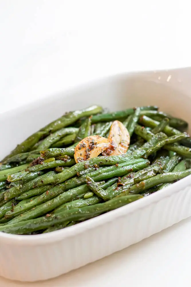 A white serving dish filled with sautéed fresh green beans, garnished with roasted garlic slices.