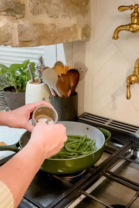 A hand adding seasoning to fresh green beans in a pan on the stovetop