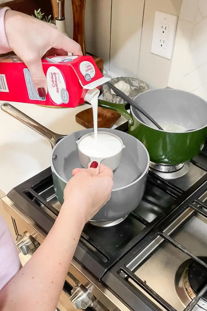 Heavy cream being measured and poured from a carton into a pot on the stove, part of the garlic mashed potato recipe.