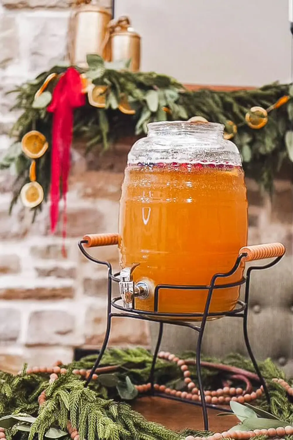 Glass drink dispenser filled with amber holiday party punch recipe on a rustic table with Christmas greenery garland and dried orange slices in the background.
