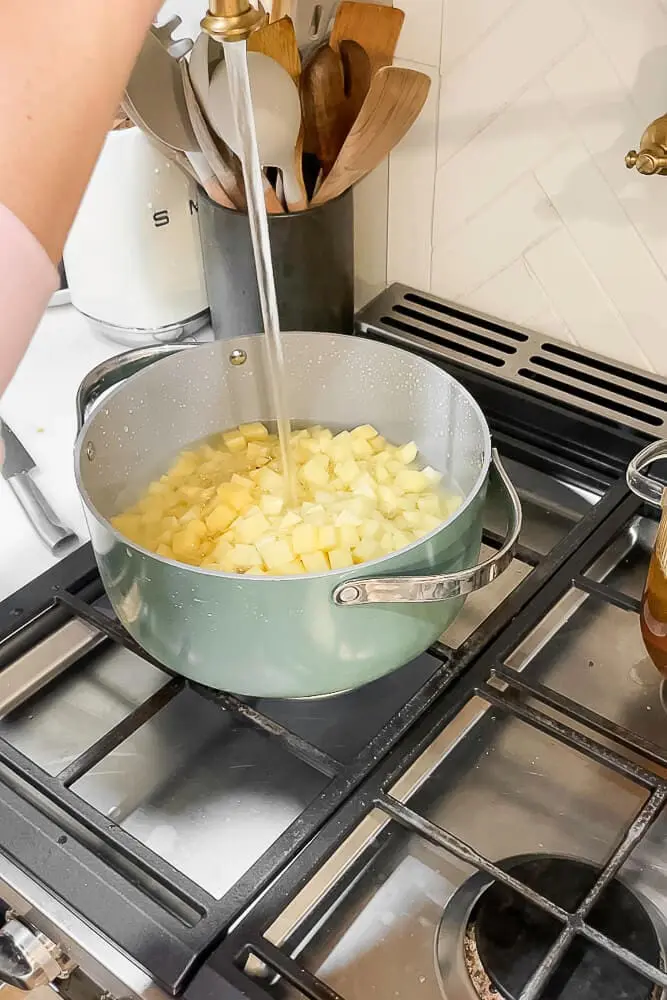 A pot filled with diced potatoes being covered with water from the faucet, preparing to boil for garlic mashed potatoes.