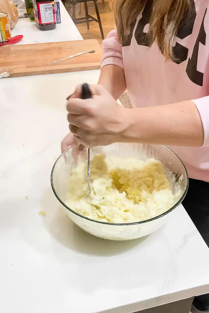 A person mashing boiled potatoes in a glass bowl with a potato masher, making creamy garlic mashed potatoes