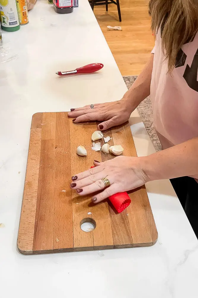 A person rolling garlic cloves in a red silicone garlic peeler on a wooden cutting board to remove the skins.