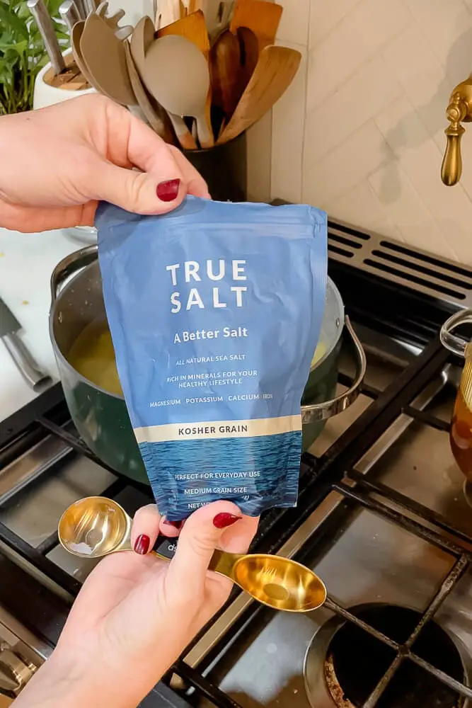 A person holding a packet of True Salt and gold measuring spoons, about to season the boiling potatoes for garlic mashed potatoes.