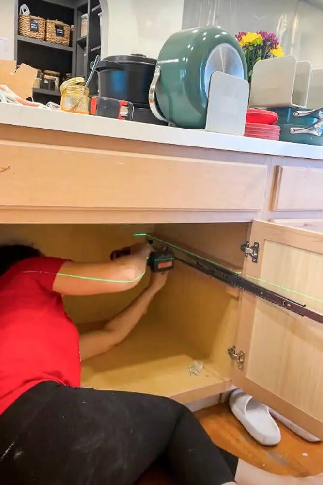 Installing a drawer slide inside a kitchen cabinet for custom pull-out shelves using a drill.