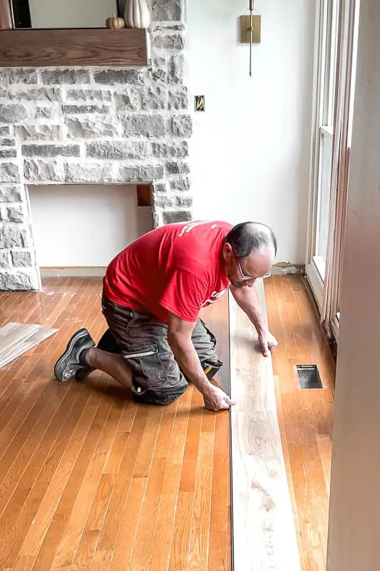 Man in red shirt installing floating plank flooring over old hardwood near a stone fireplace