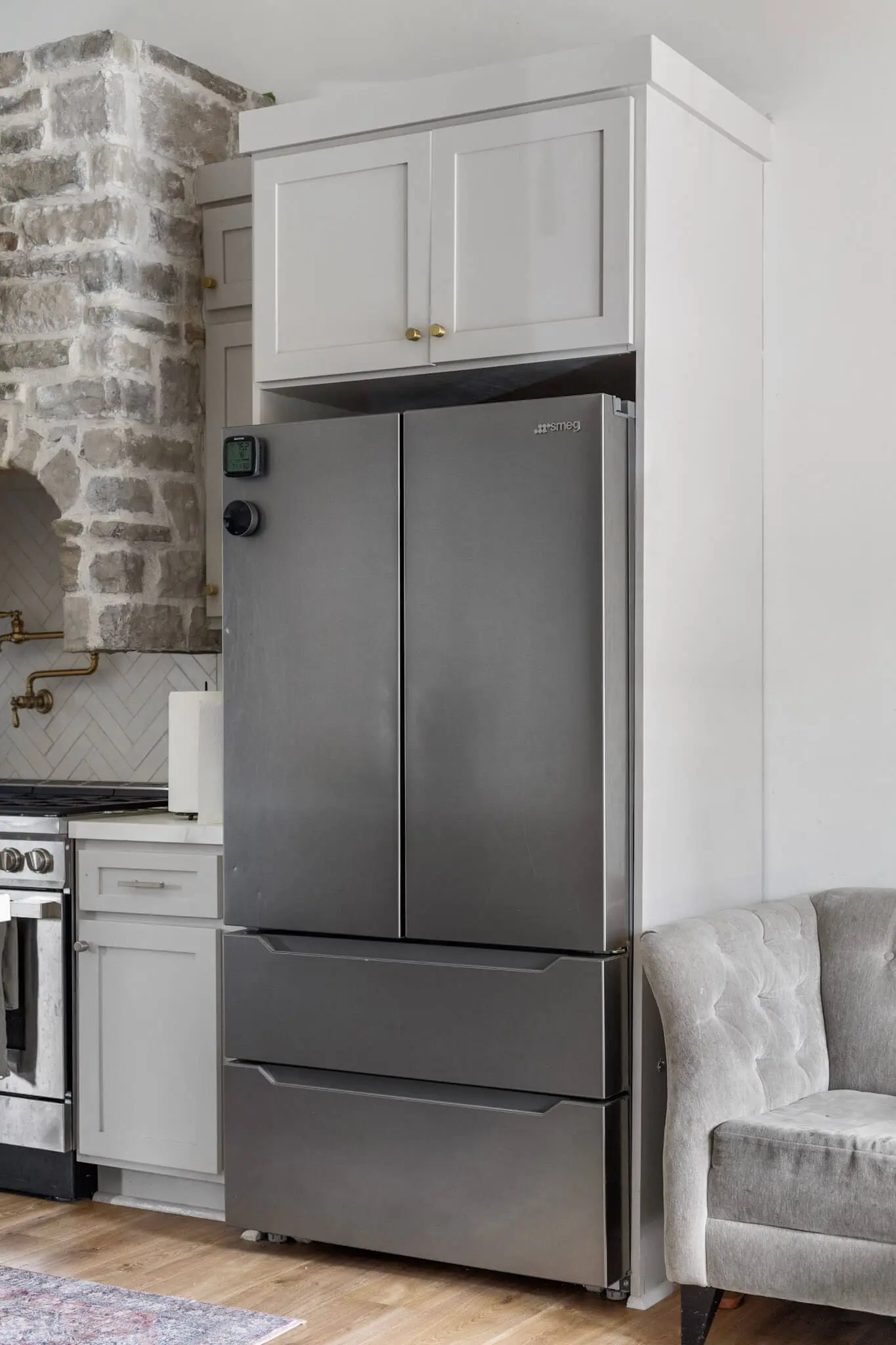 A modern stainless steel refrigerator set between white cabinets and a stone feature wall, with a tufted armchair placed nearby.