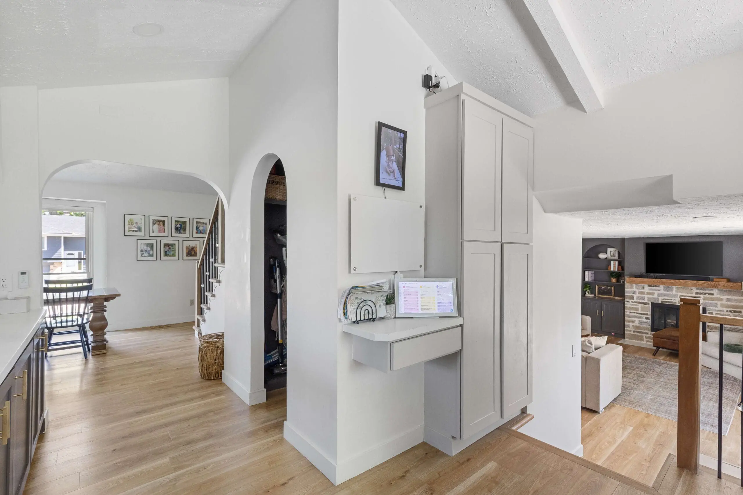 View of a small floating desk station with a digital calendar and wall-mounted planner, seamlessly integrated into a kitchen hallway near a staircase and arched doorway.