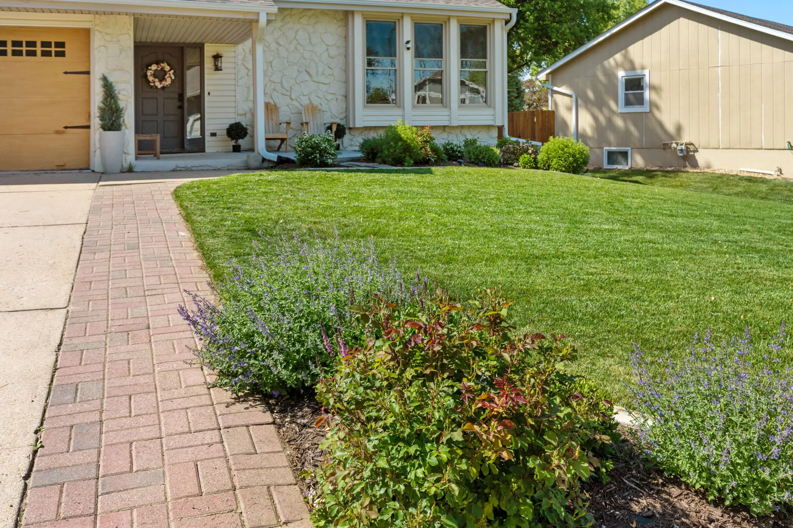 Brick walkway and fresh front yard landscaping with vibrant perennials and manicured lawn, showing the DIY paver project extending the driveway and porch entry.