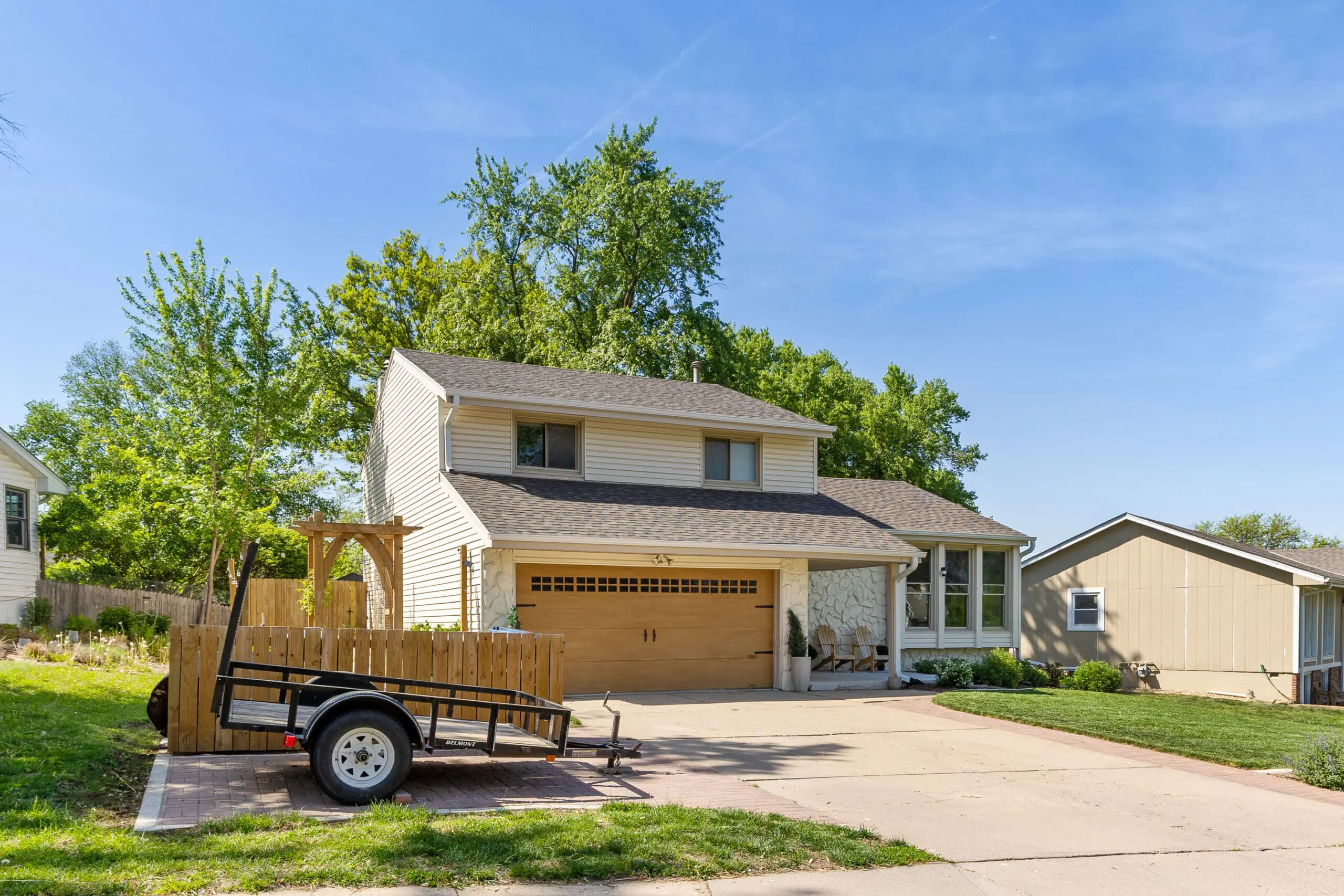 Full view of a two-story home with a DIY trash can enclosure, garden arbor, and faux wood garage door—featuring fresh landscaping and a paver pad trailer parking area.