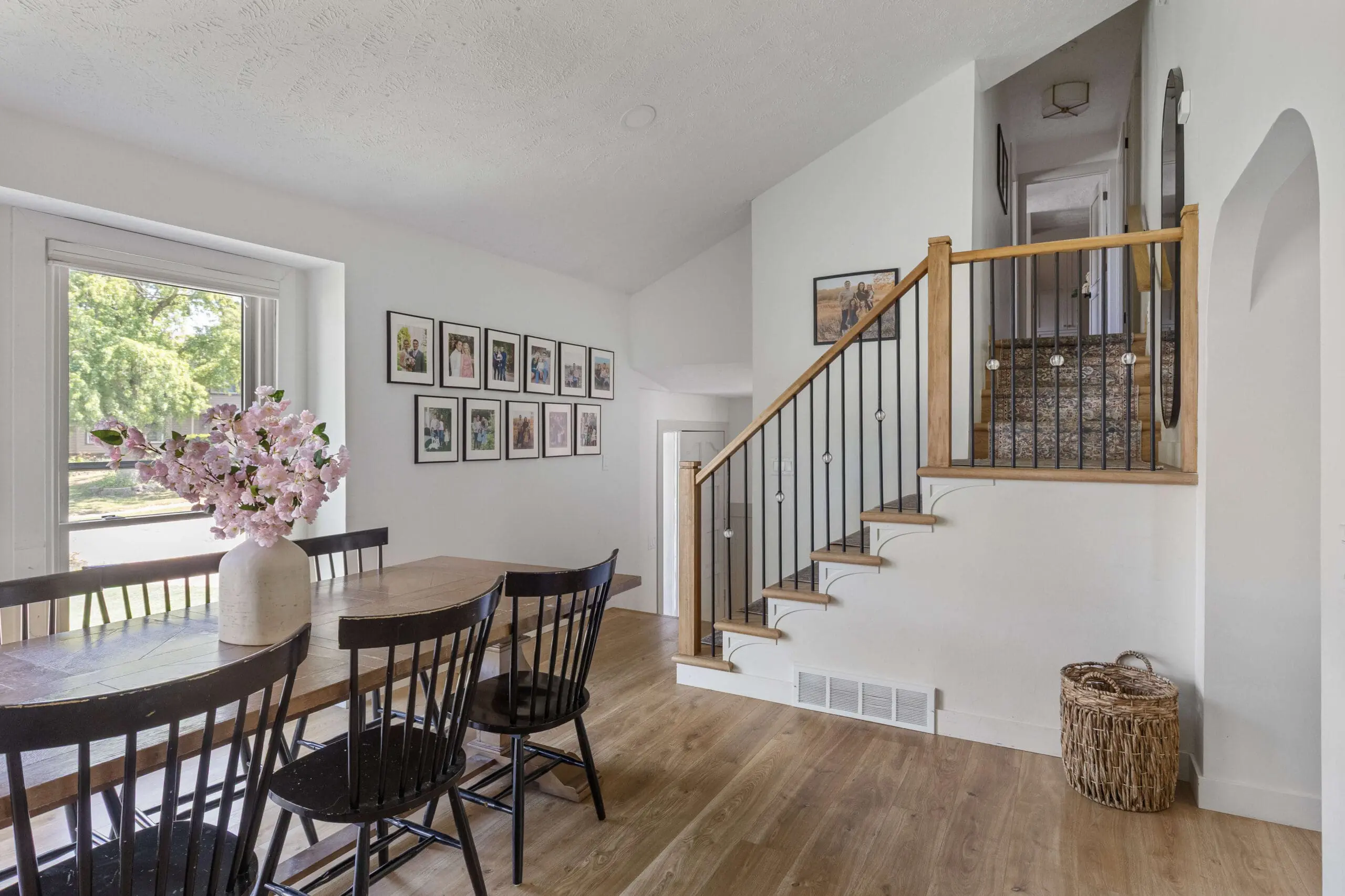 Bright dining area with a rustic wood table, black spindle chairs, and a large family photo gallery wall leading into a stairway with wood and wrought iron railing.