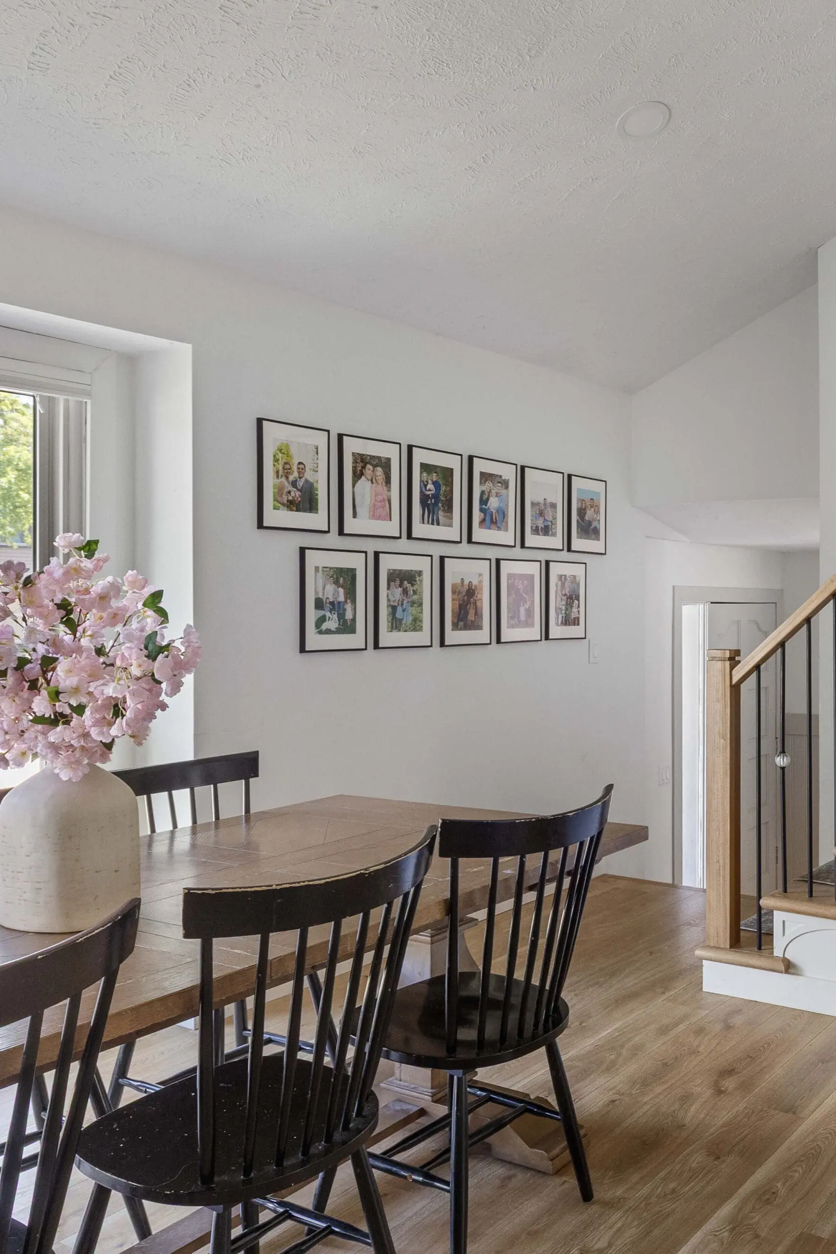 Bright dining room with a rustic wood table, black spindle-back chairs, and a symmetrical photo gallery wall featuring framed family portraits in a clean, modern layout.