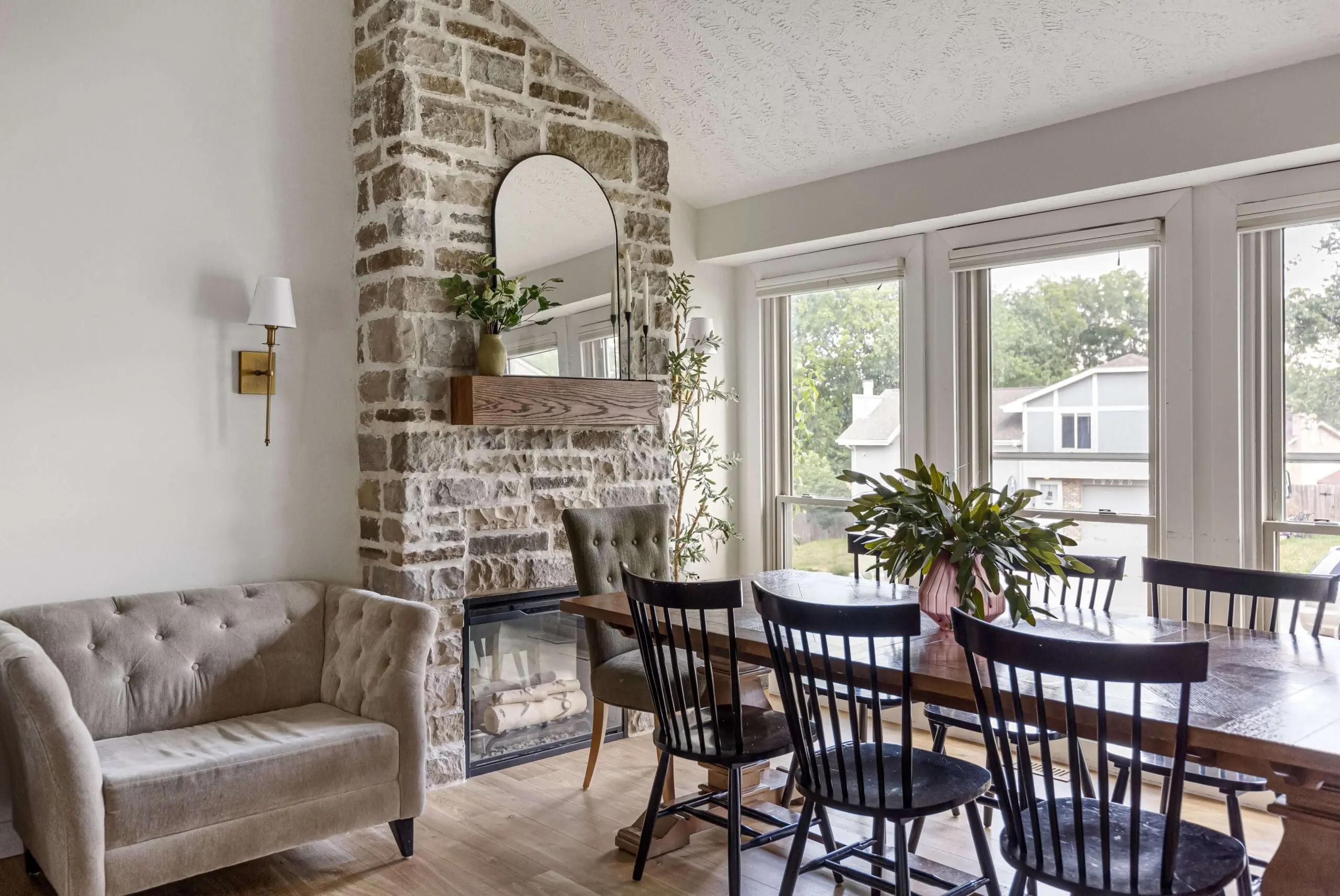 Cozy dining room featuring a DIY floor-to-ceiling stone fireplace with a wood mantel and mirror, surrounded by large windows with natural light, a tufted beige loveseat, and a wooden dining table with black spindle-back chairs.