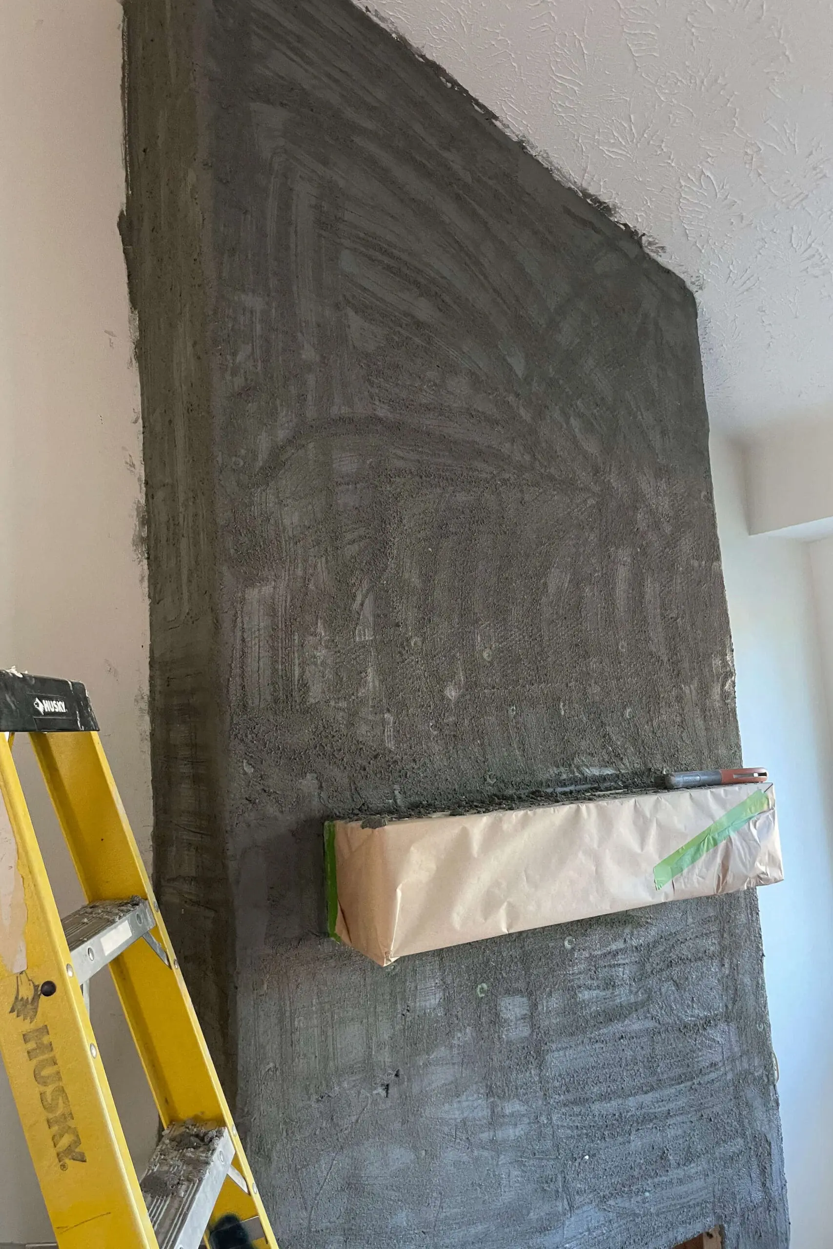 Wide shot of a living room wall covered in a dark scratch coat, with a covered mantel and yellow ladder nearby.