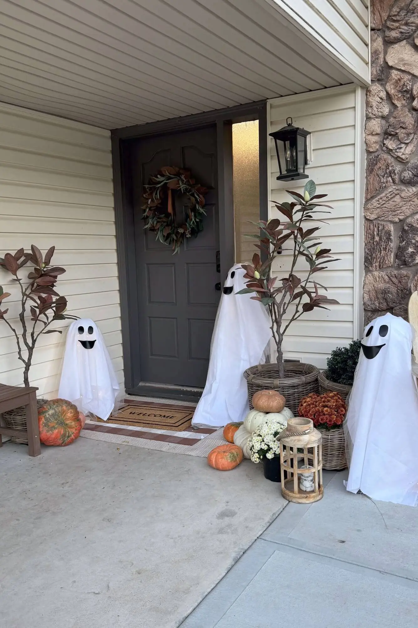 Three sheet ghosts around the front door with pumpkins, mums, a welcome mat, and a wooden lantern; daytime porch view.