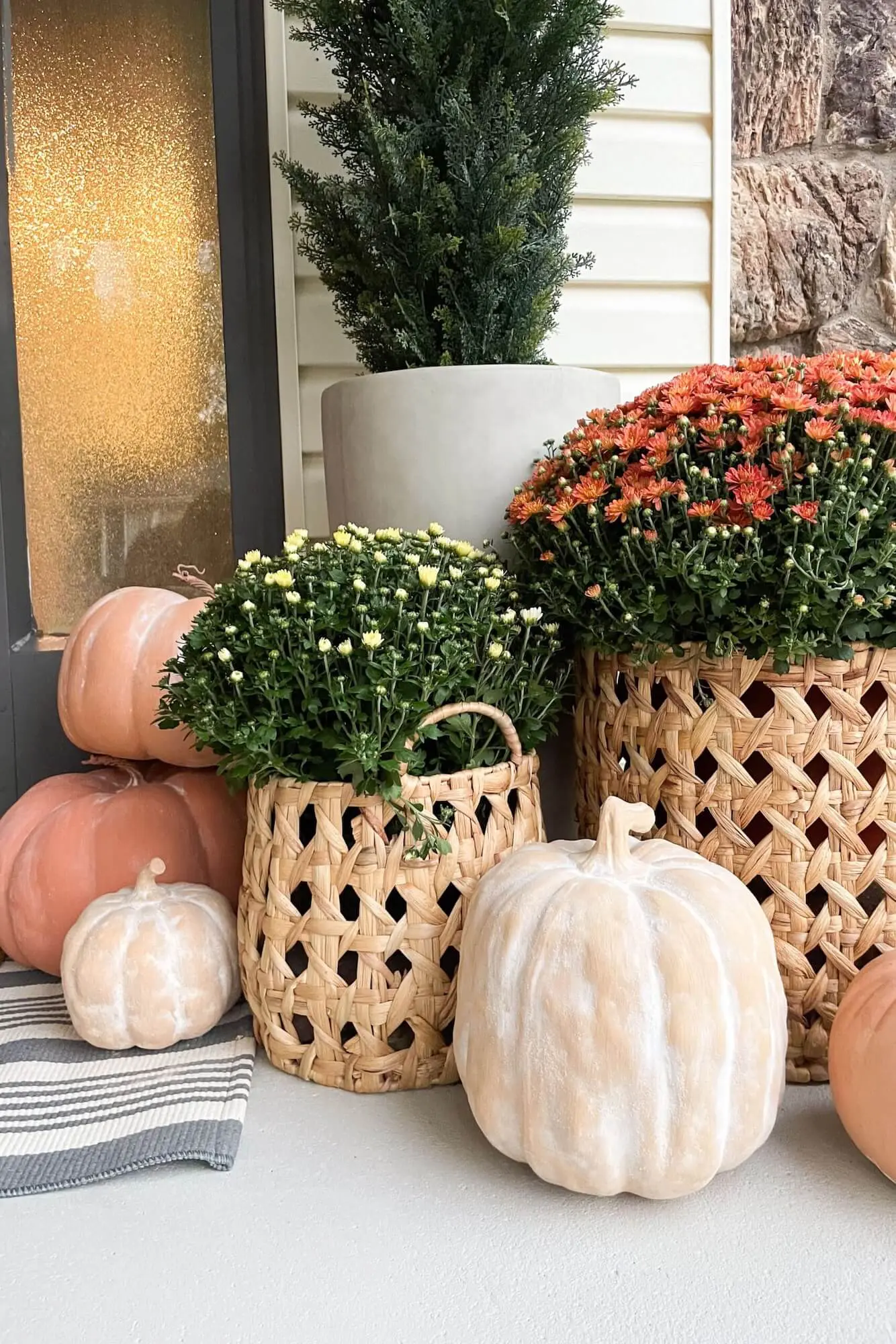 Neutral fall porch styling with woven baskets of mums, stacked pumpkins, and a tall evergreen by the front door.