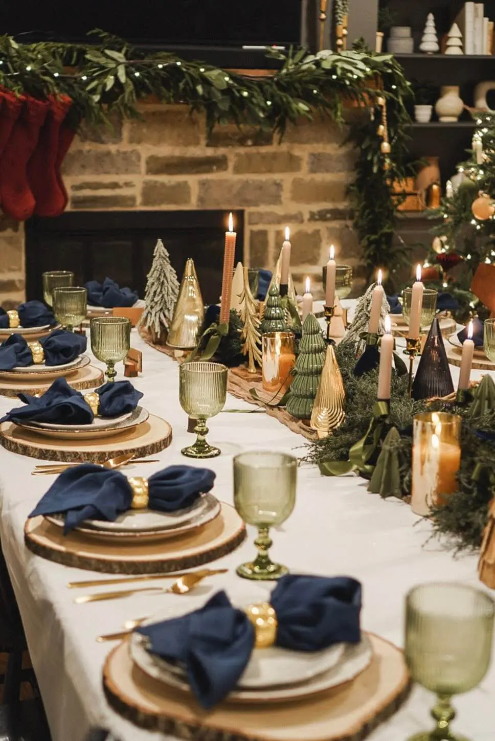 Elegant Christmas party dining table set for hosting a festive gathering, featuring natural wood slice chargers, navy napkins with gold rings, and vintage green glass goblets. The centerpiece includes miniature Christmas trees, candle-lit accents, and lush greenery, all against a warmly decorated background with a Christmas tree and garland-lit fireplace.