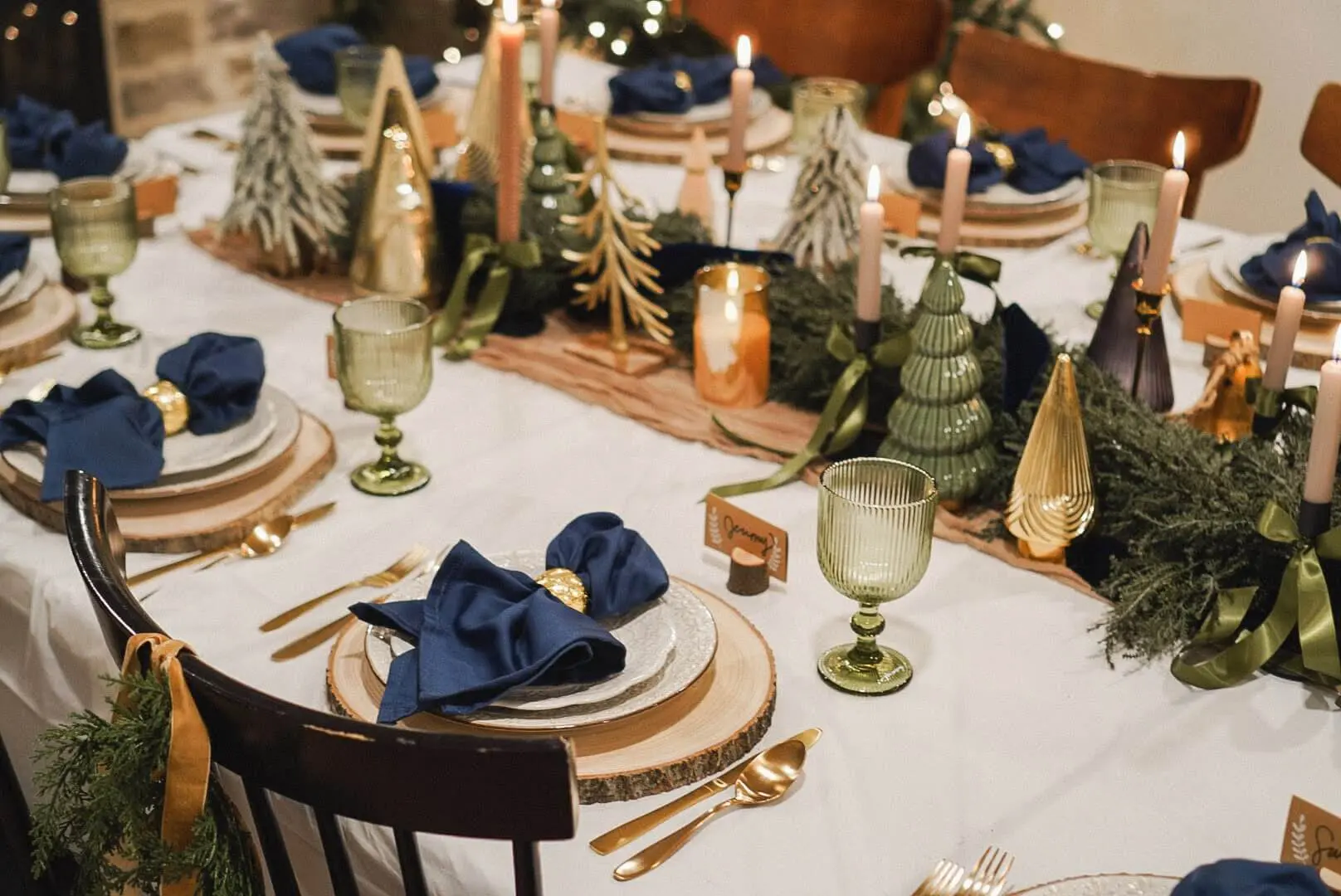 Festive tablescape perfect for hosting a Christmas party, featuring wood slice plates, dark blue napkins, and golden flatware arranged with rustic elegance. The table is adorned with candle-lit evergreen garlands, small Christmas tree figurines, and warm holiday accents, creating a cozy yet refined ambiance for holiday gatherings.