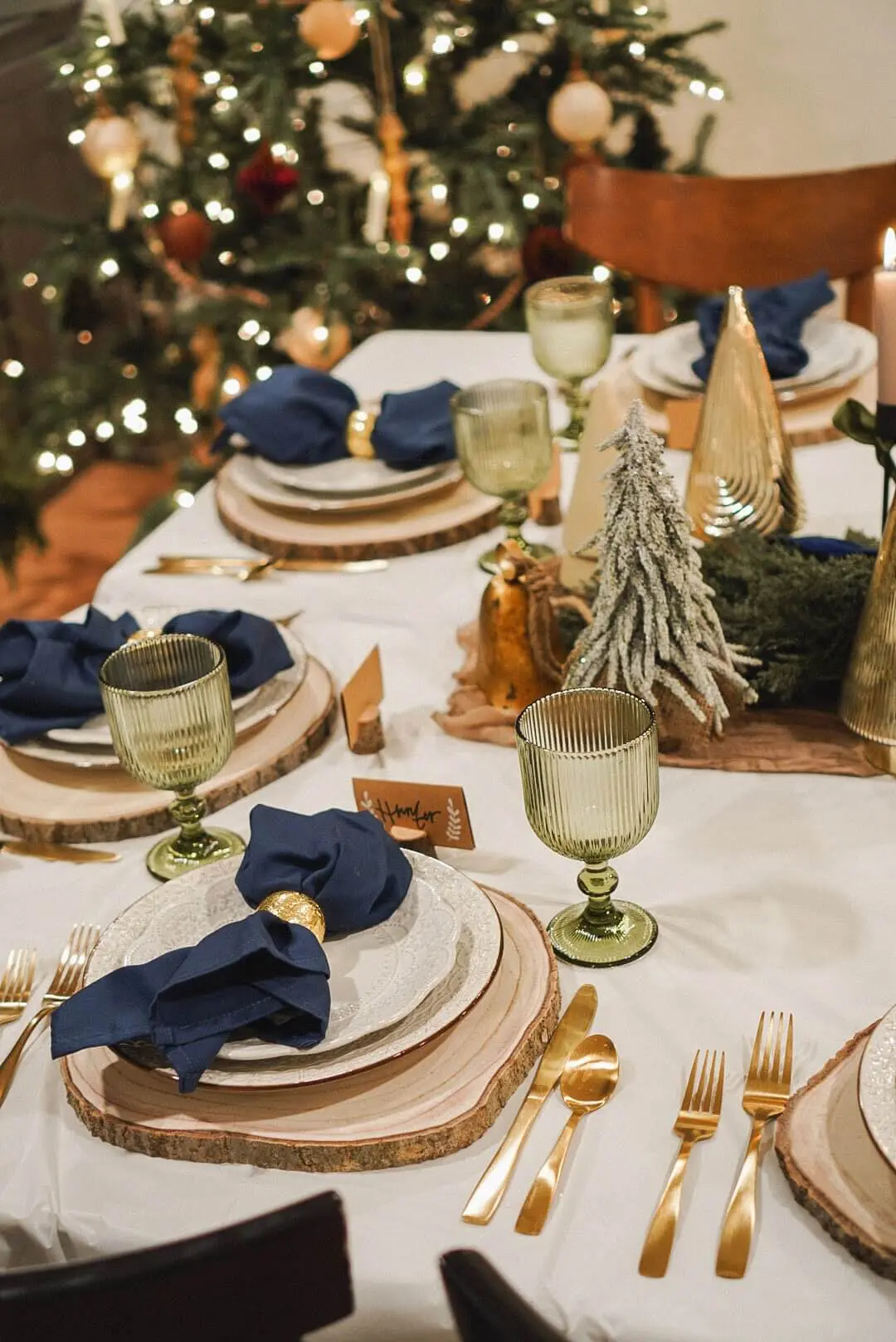 Close-up of a cozy Christmas dinner setup, ideal for hosting a holiday party, showing wood slice plates, navy blue napkins with golden napkin rings, and sage-green goblets. Decor includes candlelight, miniature frosted pine trees, and gold holiday accents, creating an inviting festive table setting with a warmly lit Christmas tree nearby.