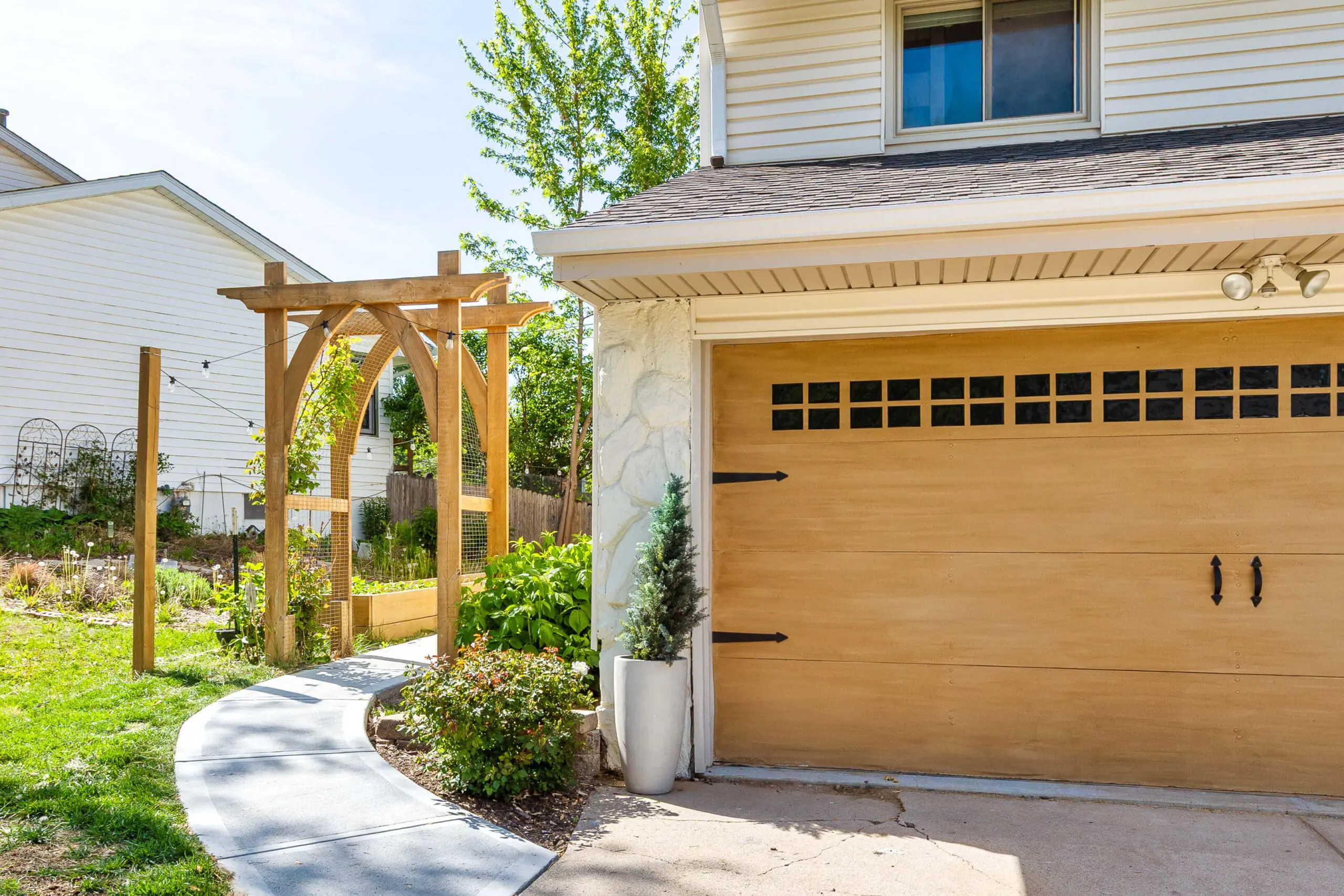 Faux wood garage door next to a curved sidewalk leading to a DIY wooden arbor with wire trellis and backyard garden beds.