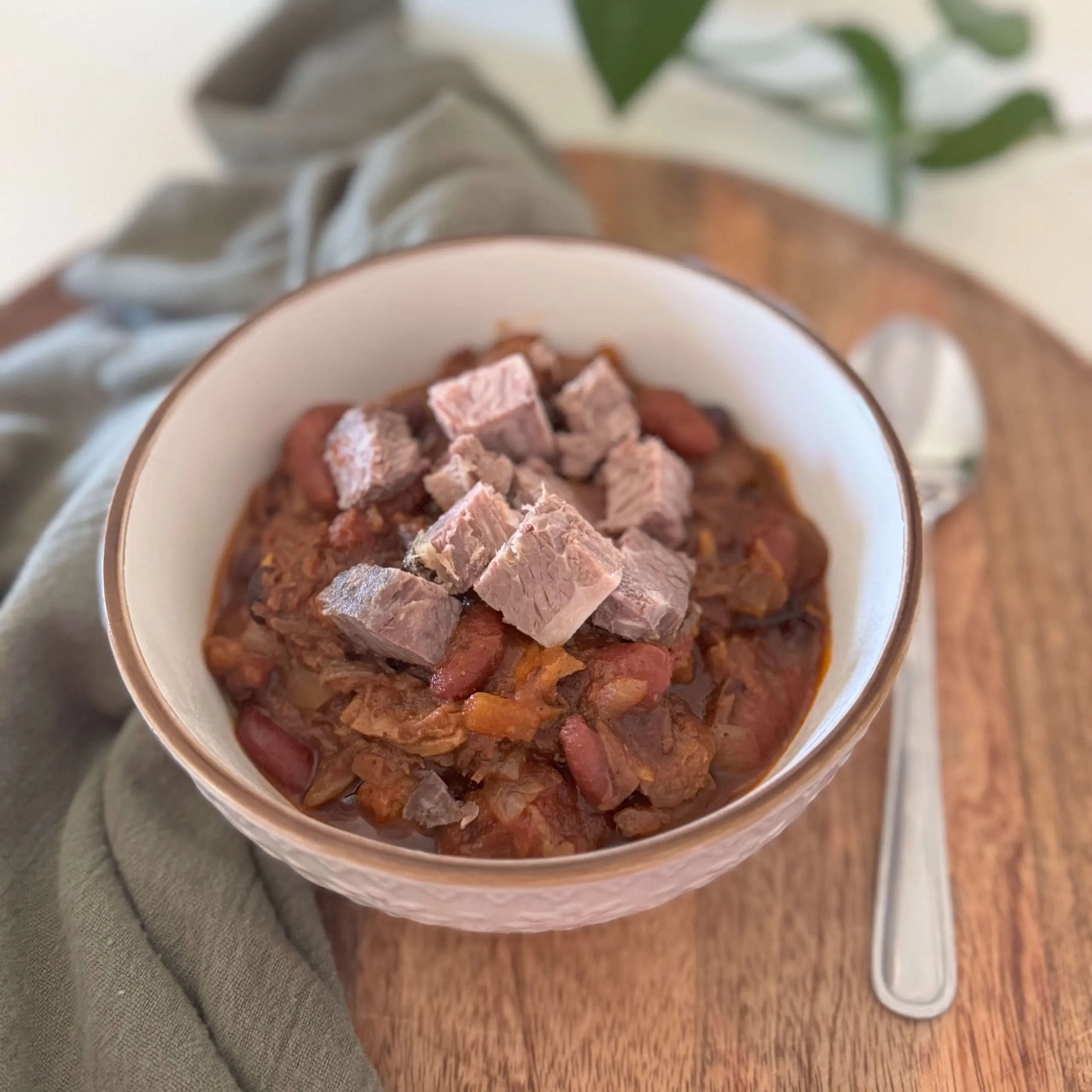 A bowl of hearty brisket chili topped with tender cubes of smoked brisket, served on a wooden tray with a spoon and green linen napkin—perfect for a cozy brisket chili recipe.