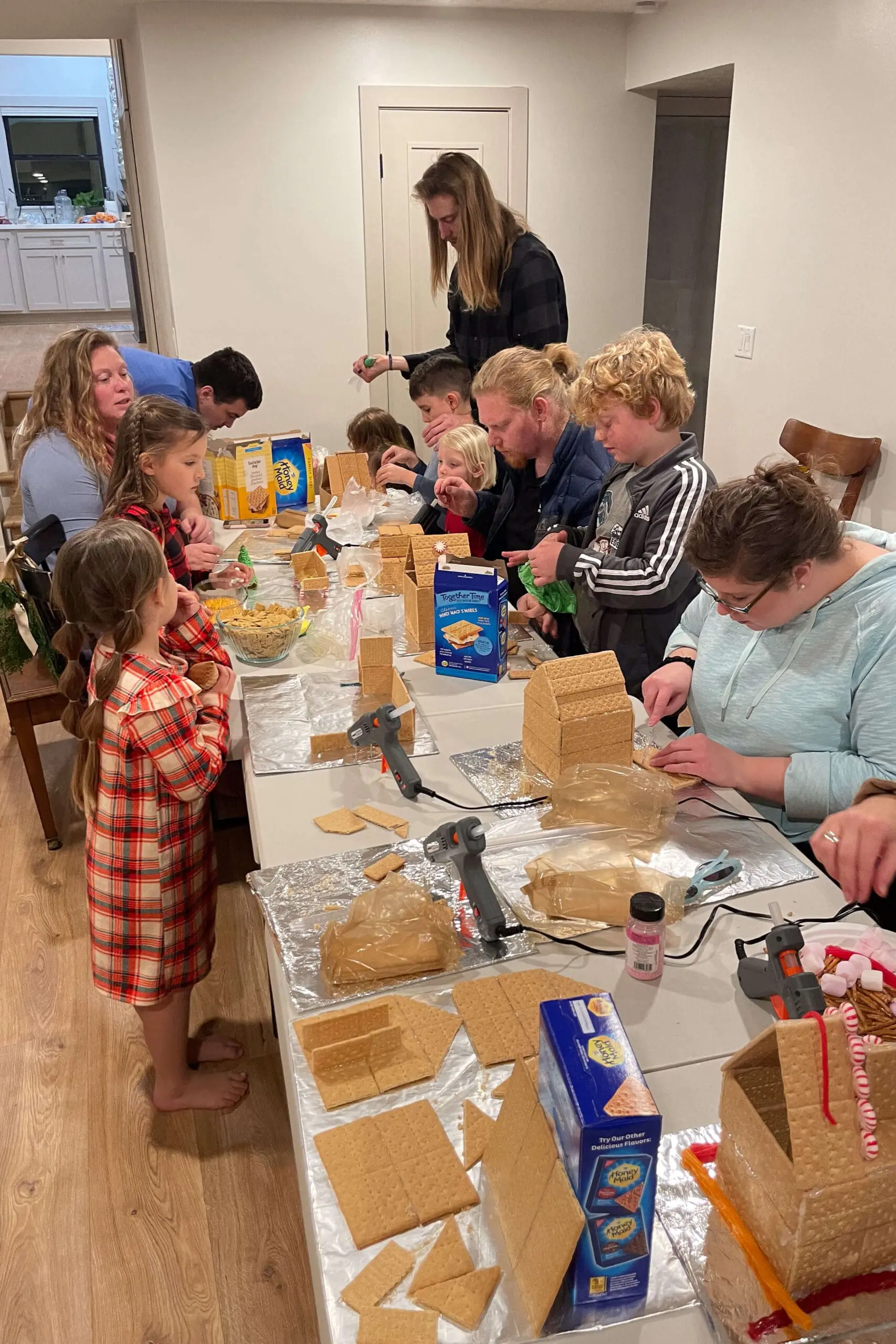 Family gathered around a table constructing gingerbread houses, each focused on assembling graham cracker walls and roofs. This scene captures holiday fun with kids and adults alike, surrounded by graham crackers, frosting, and festive decorations.