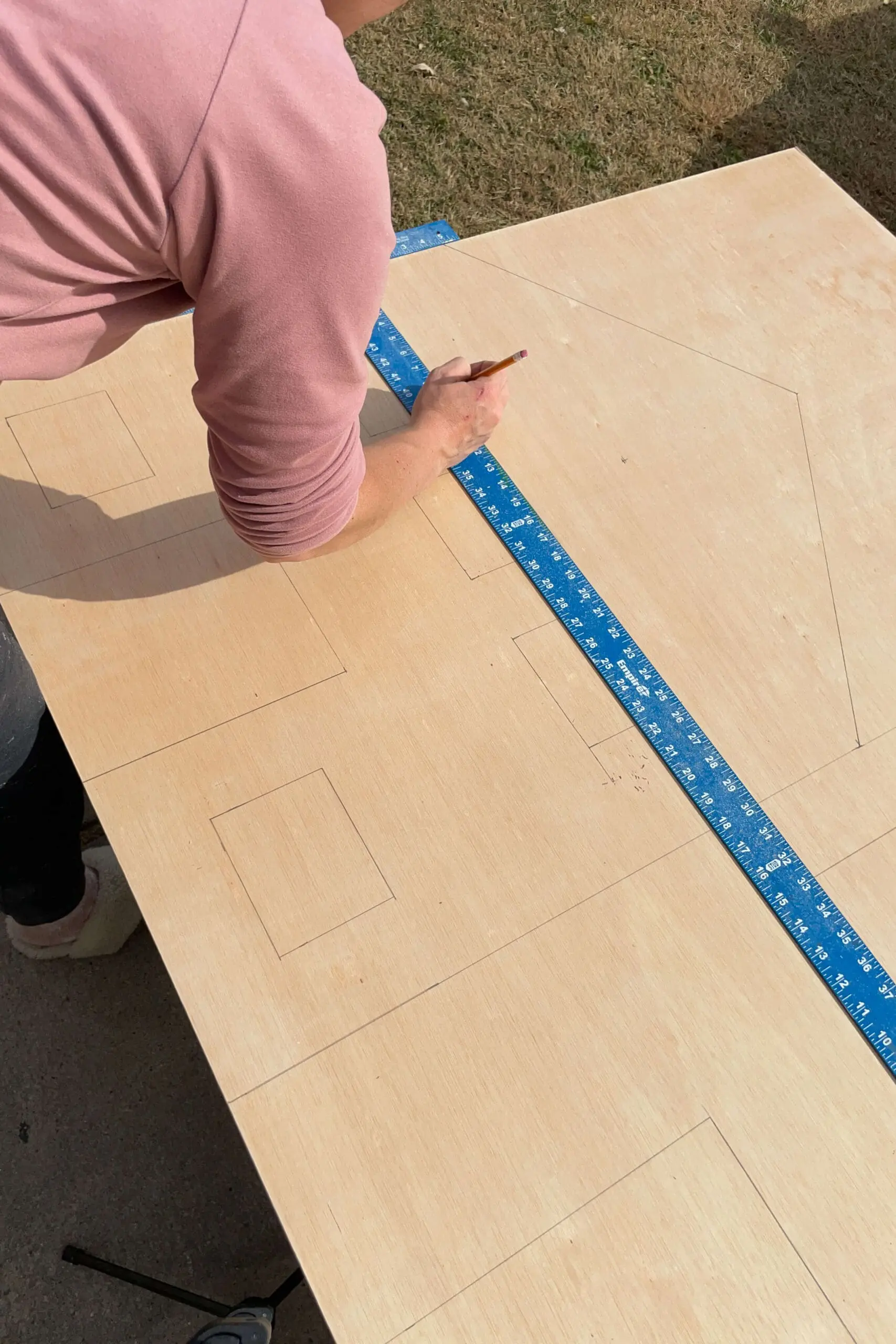 Person outlining windows and door shapes on plywood with a pencil, creating the framework for a DIY wooden gingerbread house. The precise measurements and markings bring this holiday craft project to life.