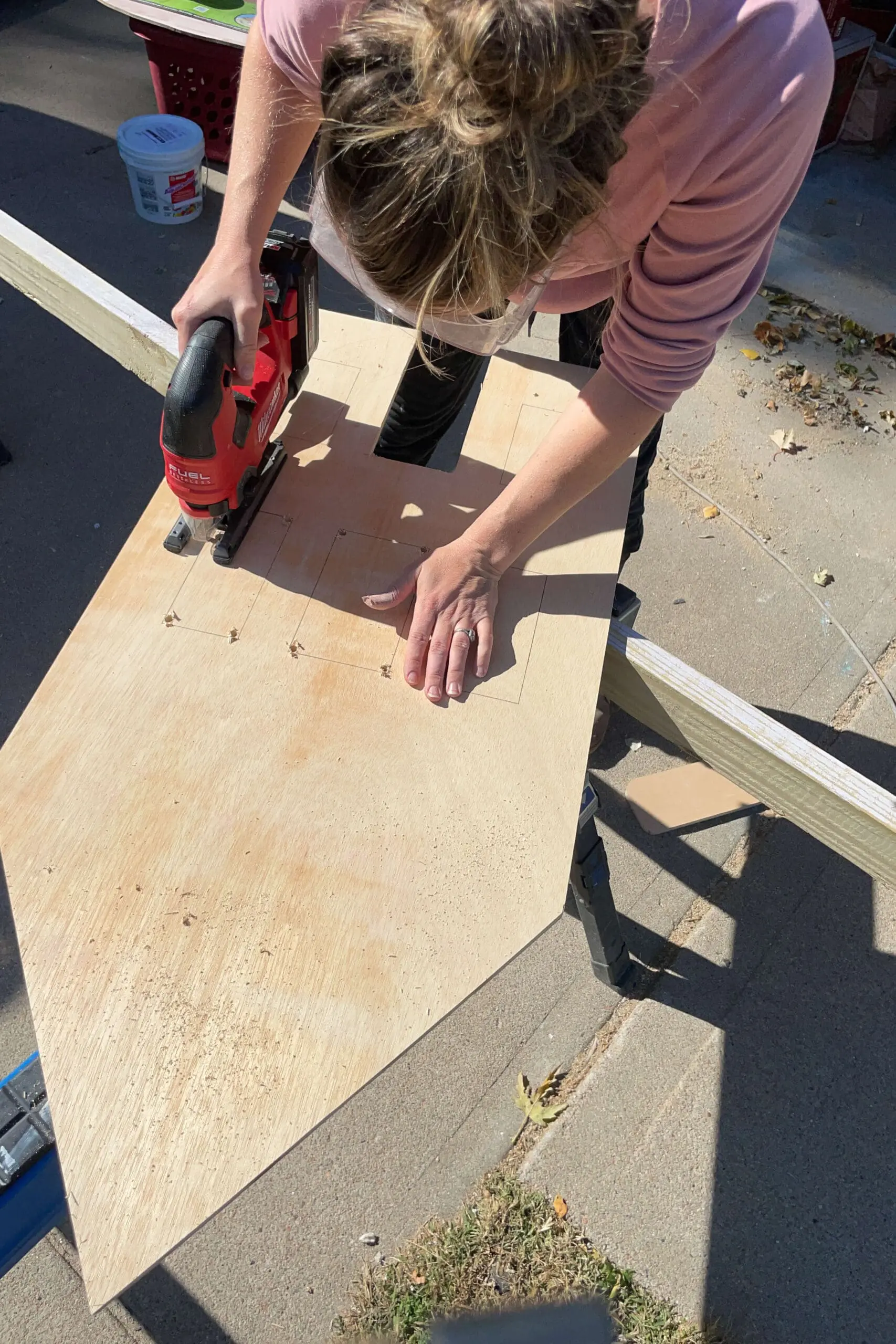 Person using a jigsaw to cut out the door and window sections of a plywood panel, assembling the structure of a wooden gingerbread house. This hands-on process showcases the attention to detail in holiday decor construction.