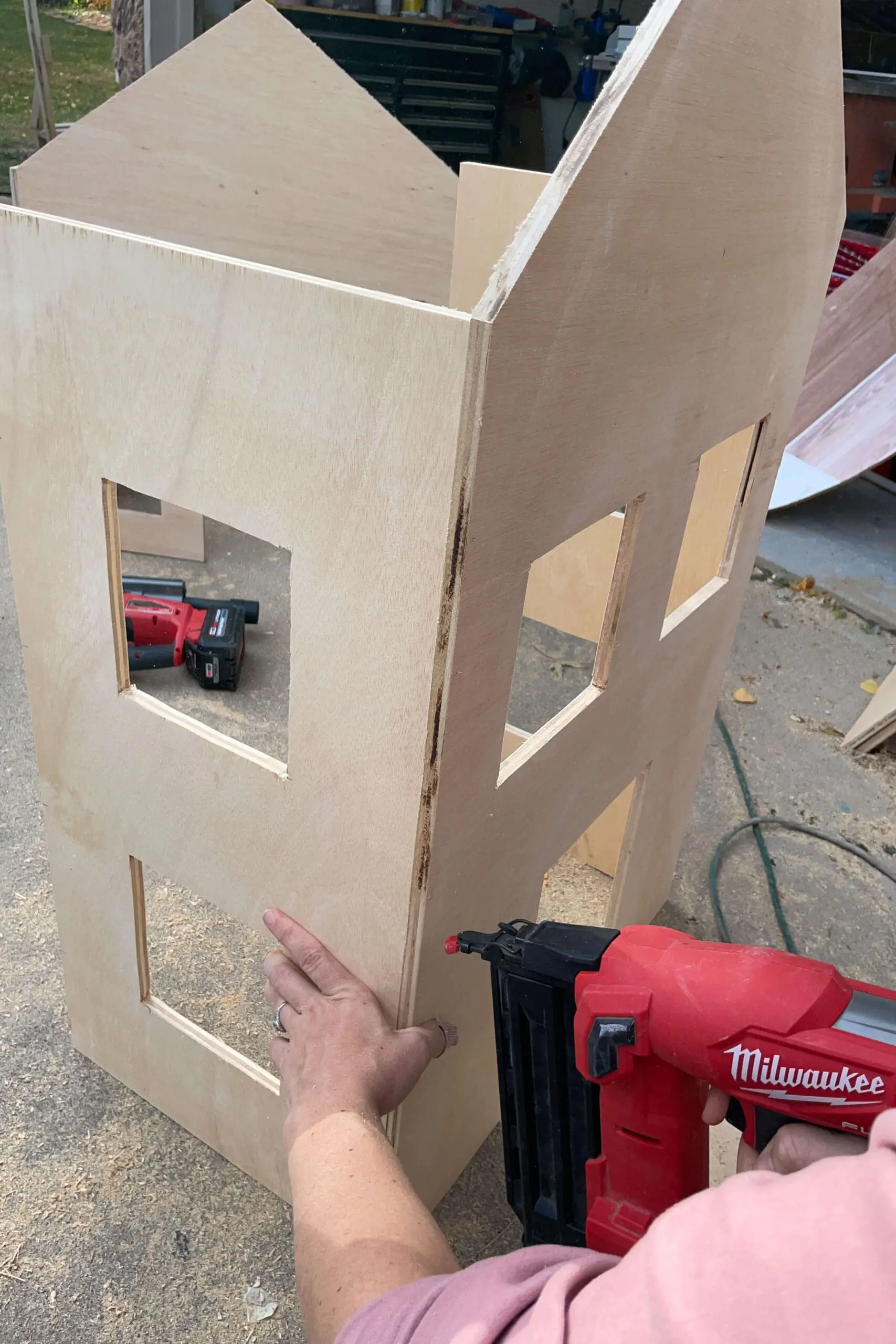 Partially assembled wooden gingerbread house structure with window cutouts, held together using a nail gun. The DIY project is beginning to take shape, showcasing a rustic, holiday-inspired decor idea.