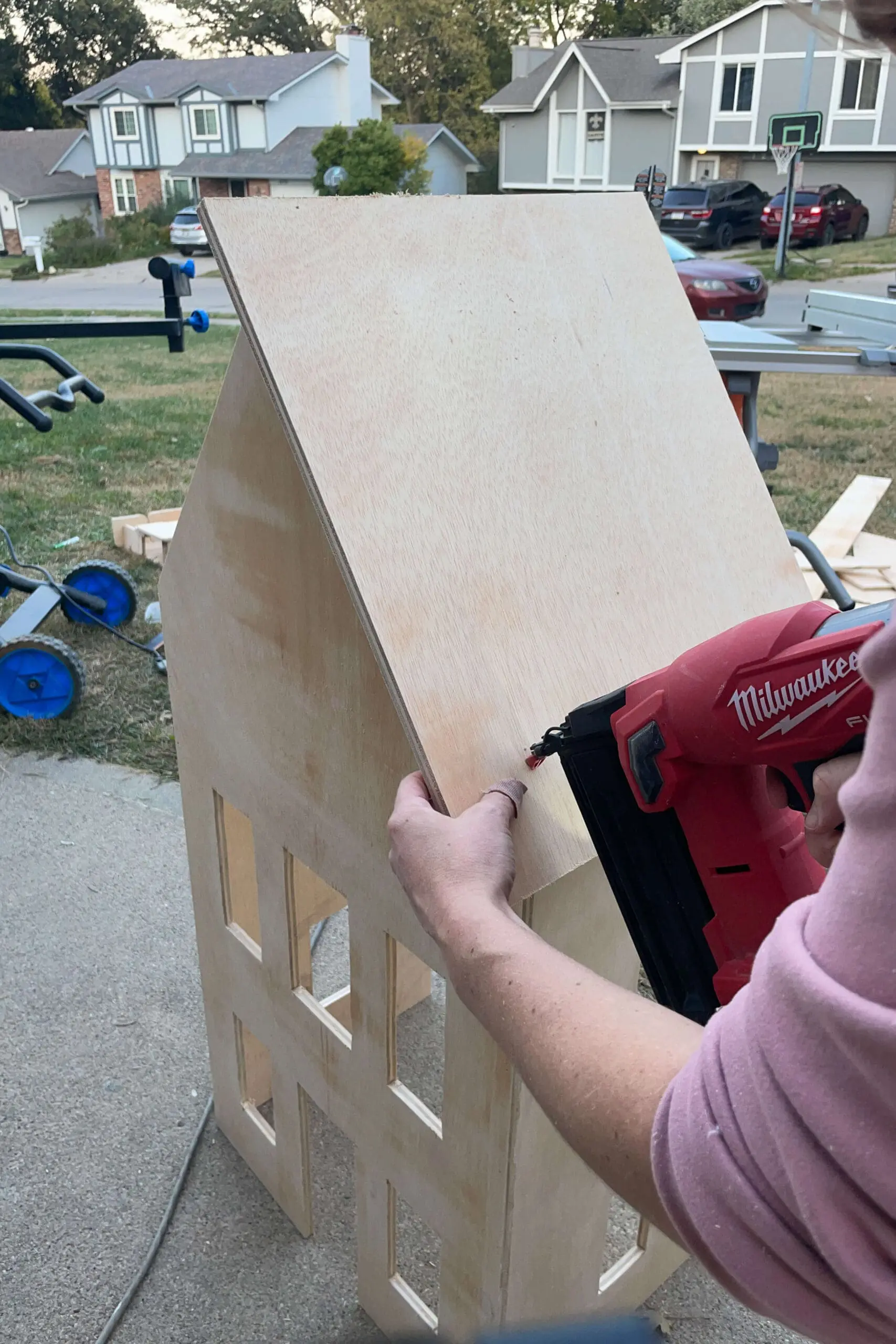 Person attaching the triangular roof panels to a wooden gingerbread house structure using a nail gun. The roof installation is a key step in this festive DIY project, giving the structure its final holiday-ready form.