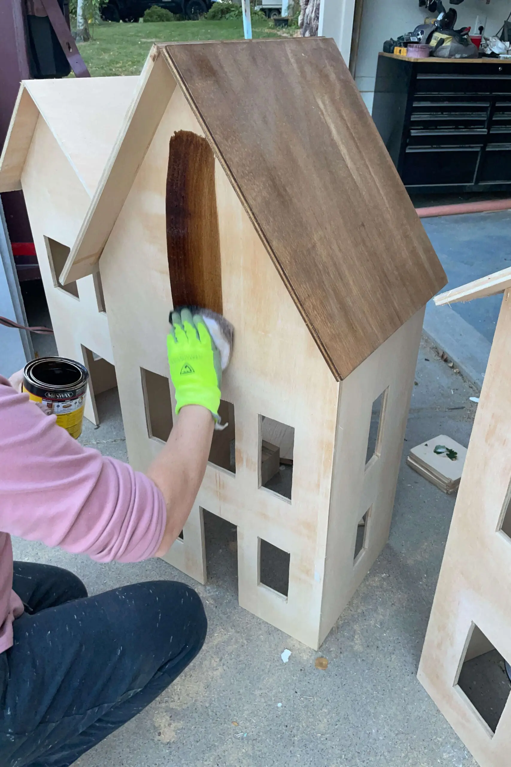 Person applying a rich brown wood stain to the roof of a wooden gingerbread house, creating a rustic look for this holiday decor piece. The stain highlights the natural wood grain, giving the structure a warm and inviting aesthetic.