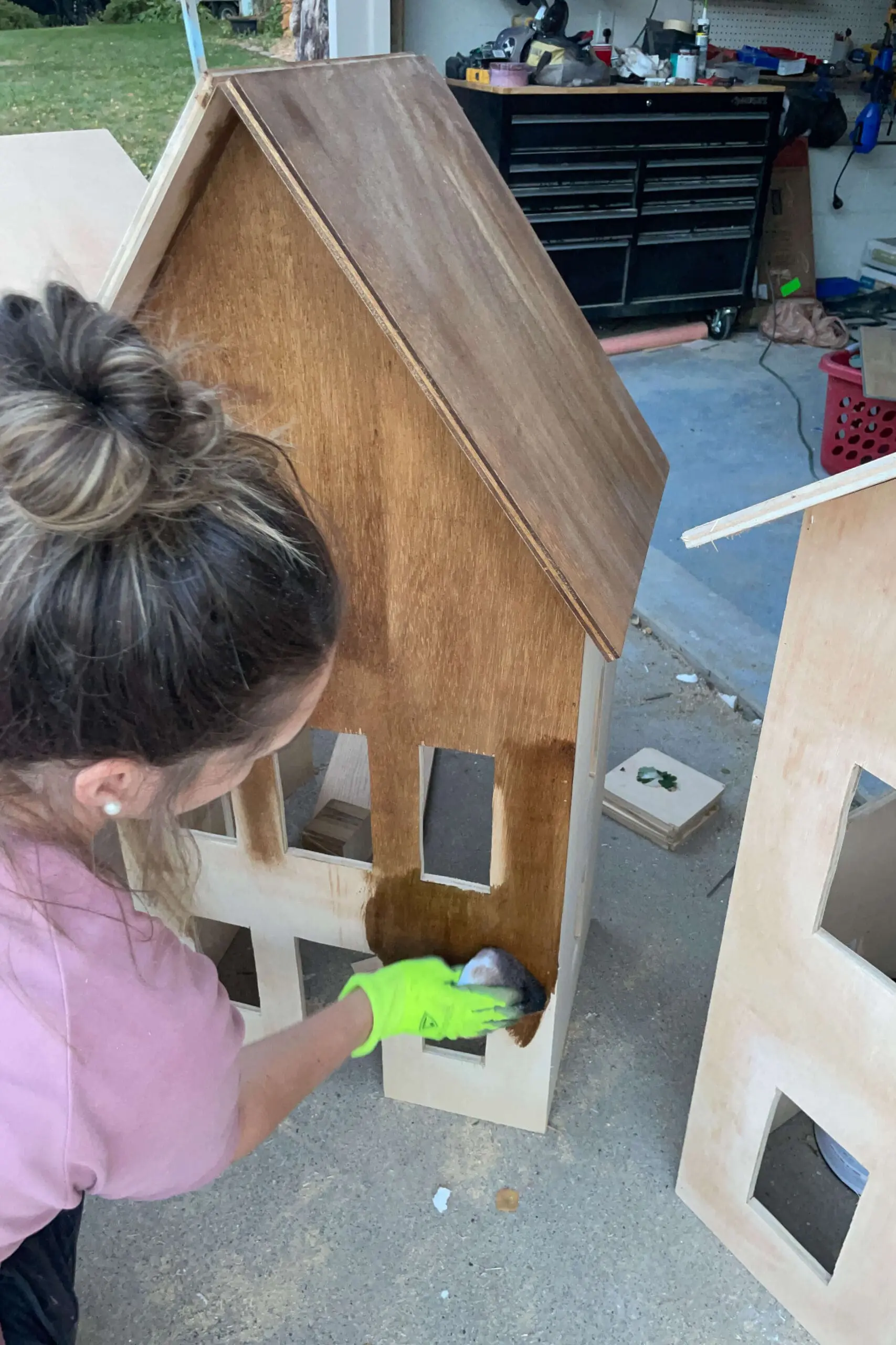 Close-up of a person staining the front panel of a wooden gingerbread house, emphasizing the rich, dark finish that brings out the charm of the wooden structure. The project showcases attention to detail, perfect for holiday decor.