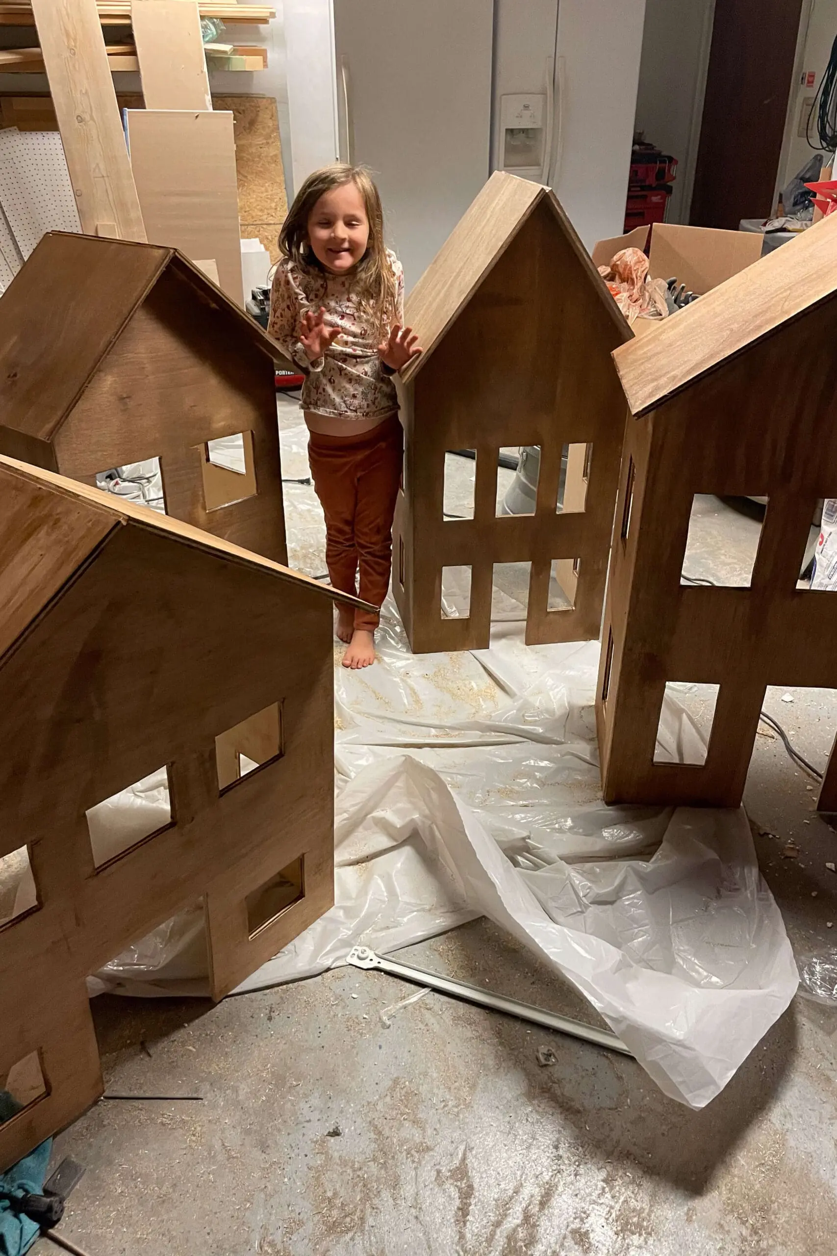 Young girl standing between several completed wooden gingerbread houses with stained exteriors. The charming village setup highlights the festive potential of these handcrafted holiday decorations.