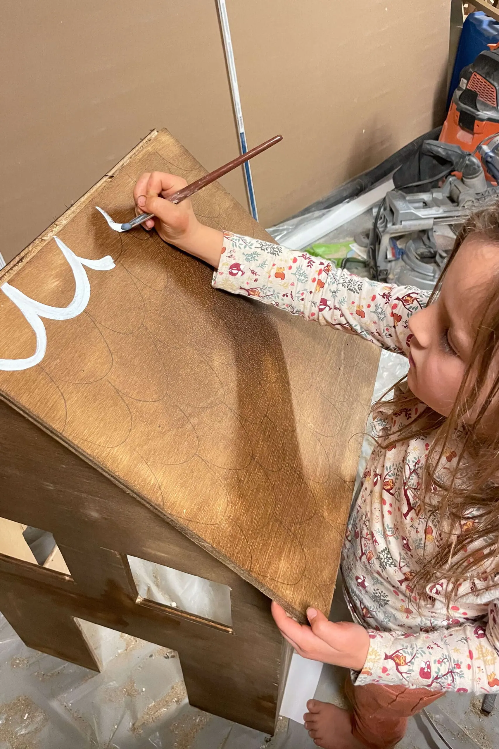 Young girl carefully painting white details on the roof of a wooden gingerbread house, adding whimsical designs that resemble frosting. This step transforms the project into a delightful holiday display with intricate, seasonal details.