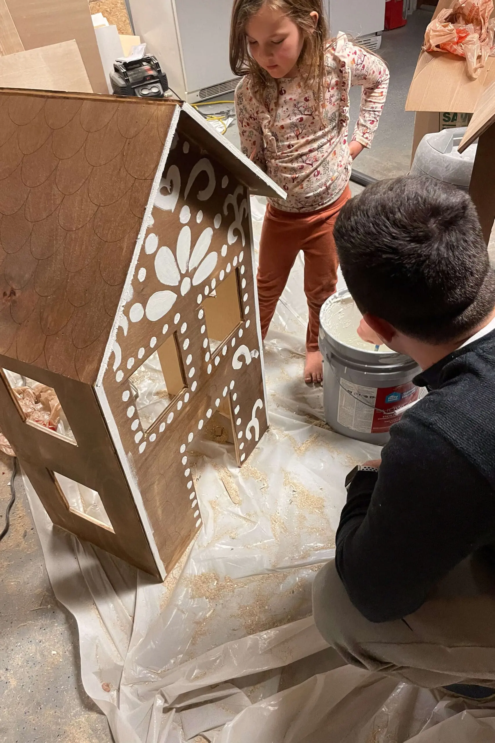 Child and adult adding white decorative paint to a stained wooden gingerbread house, creating a classic frosting effect. The intricate designs bring the holiday structure to life, capturing the festive spirit.