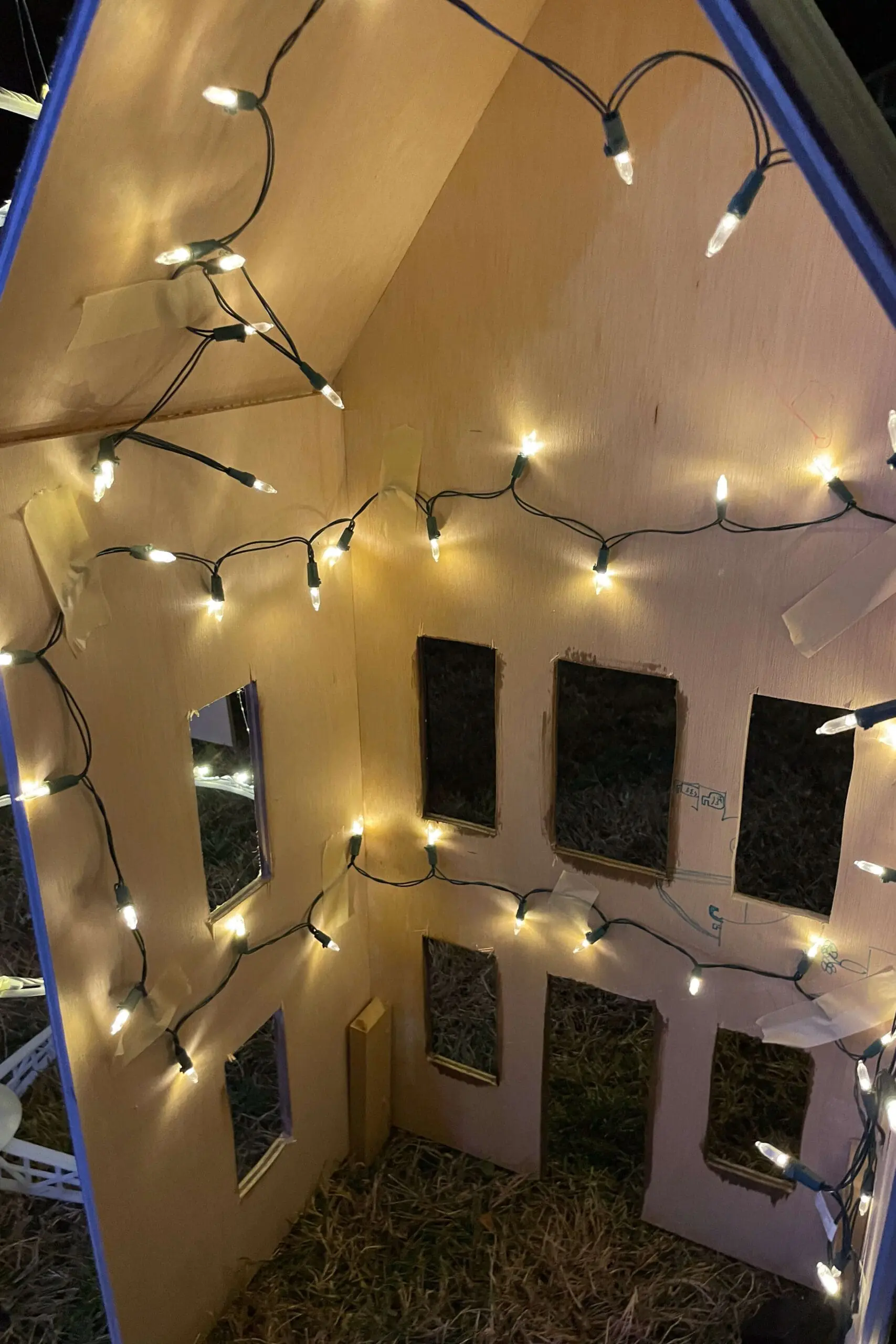 Interior view of a wooden gingerbread house illuminated with string lights, creating a cozy glow that highlights the handcrafted windows and structure. The soft lighting adds warmth, making it a magical holiday display piece.