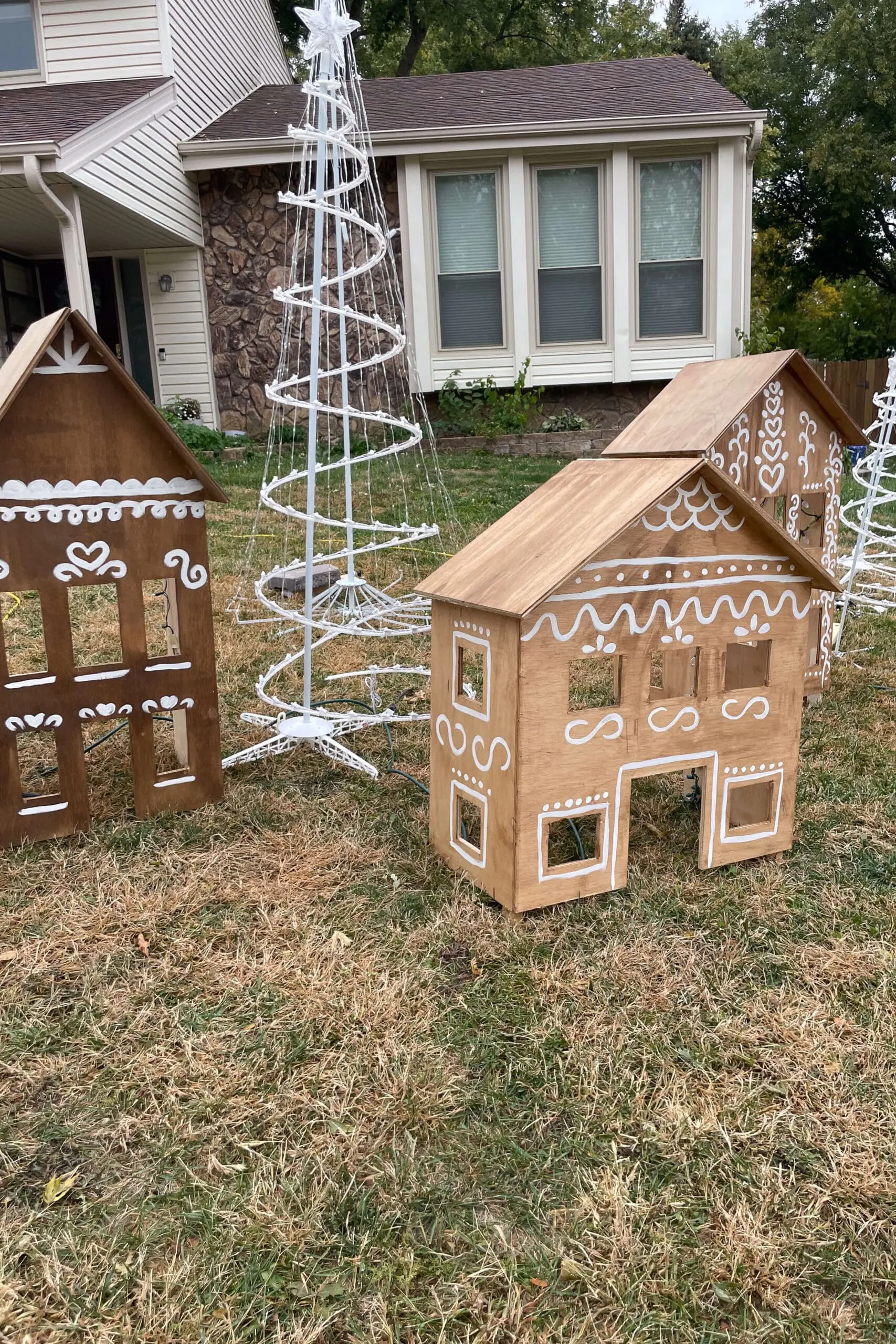 Wooden gingerbread houses with painted white details are arranged in the yard alongside tall, spiral light trees, ready to charm neighbors with a festive display.