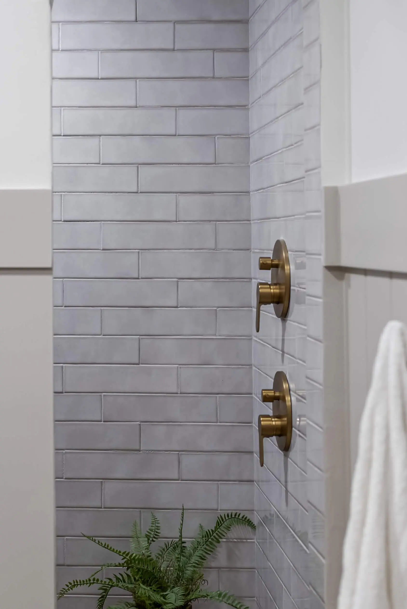 Minimalist shower corner with sleek gray subway tiles arranged in a staggered pattern, accented by two matte gold shower handles mounted on the wall, bringing a modern, sophisticated touch to the space.