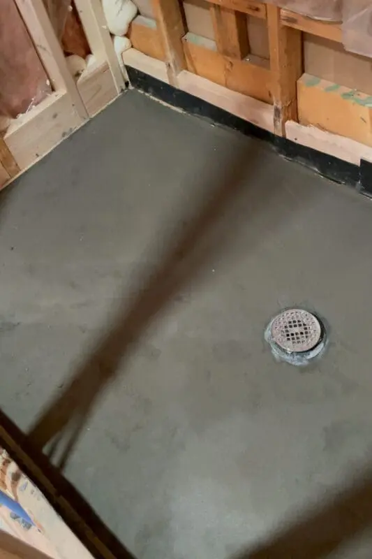 Freshly poured concrete shower pan with a centered floor drain, part of a bathroom renovation. The smooth concrete surface contrasts with the unfinished walls, which have exposed insulation and wooden framing, signaling the ongoing work in the space.